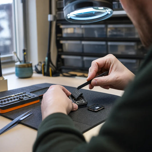 Technician holding an opened Apple Watch case with a pry tool under a magnifying lamp, the screen set aside on a repair mat